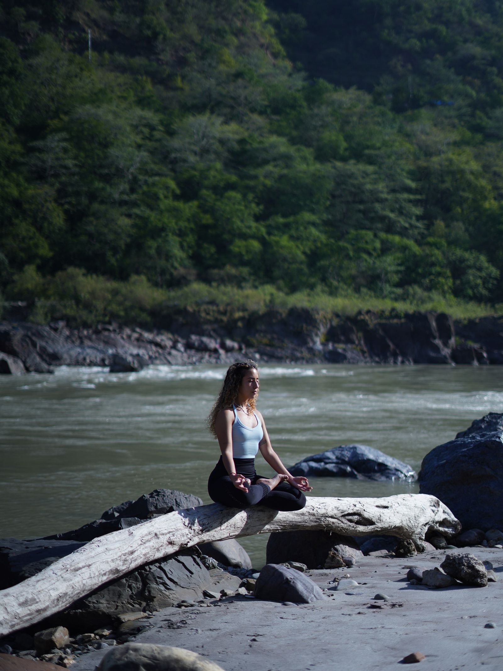 A woman practices meditation on a log by a riverside, surrounded by lush forests.