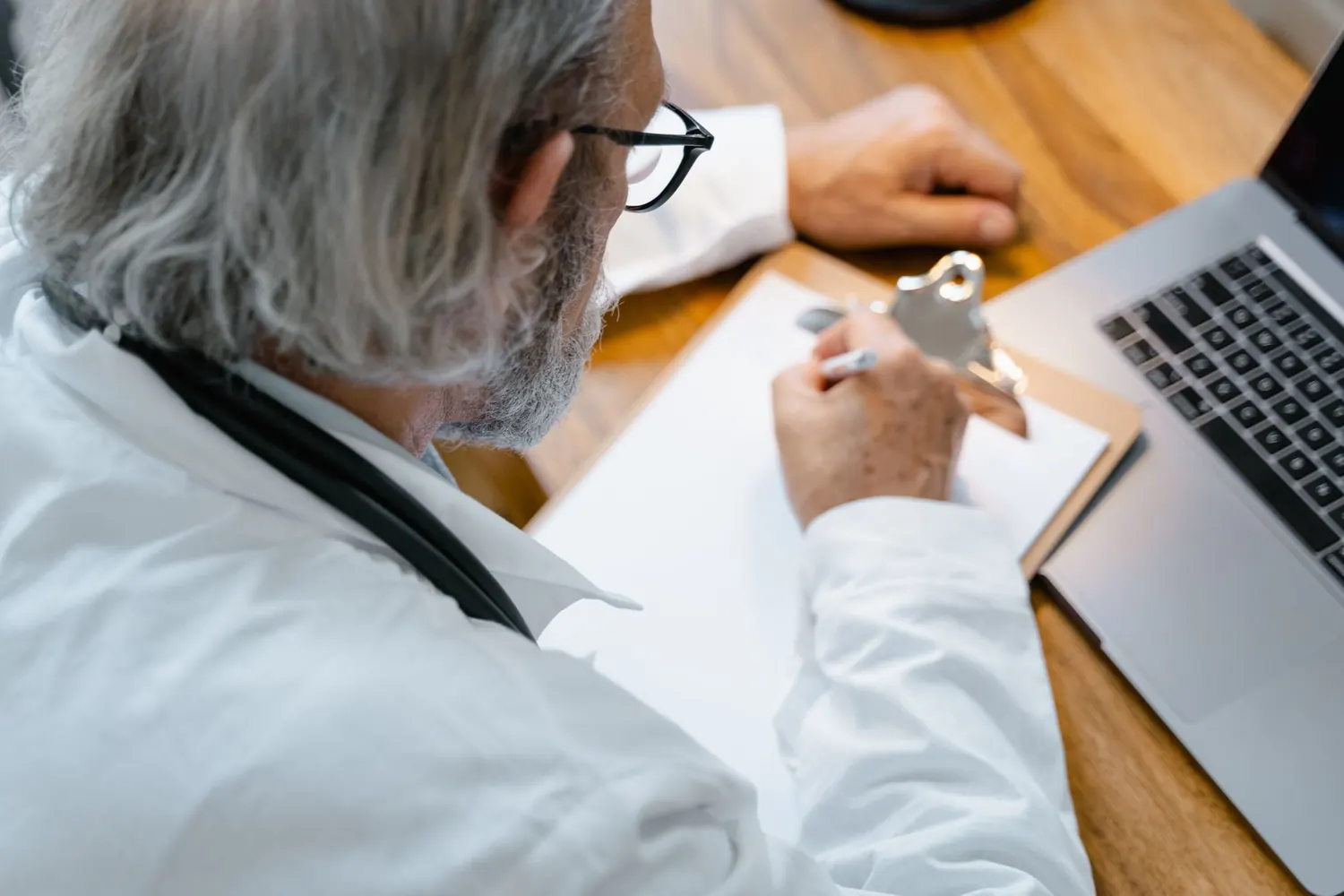 Soignant vu de dos écrivant des notes sur une planchette près d'un ordinateur portable